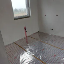 Przygotowanie do montażu ogrzewania podłogowego: mata grzewcza z folią aluminiową i czerwoną siatką na podłożu w surowym wnętrzu, widoczne wyprowadzenia instalacji wodnej i elektrycznej na ścianie.