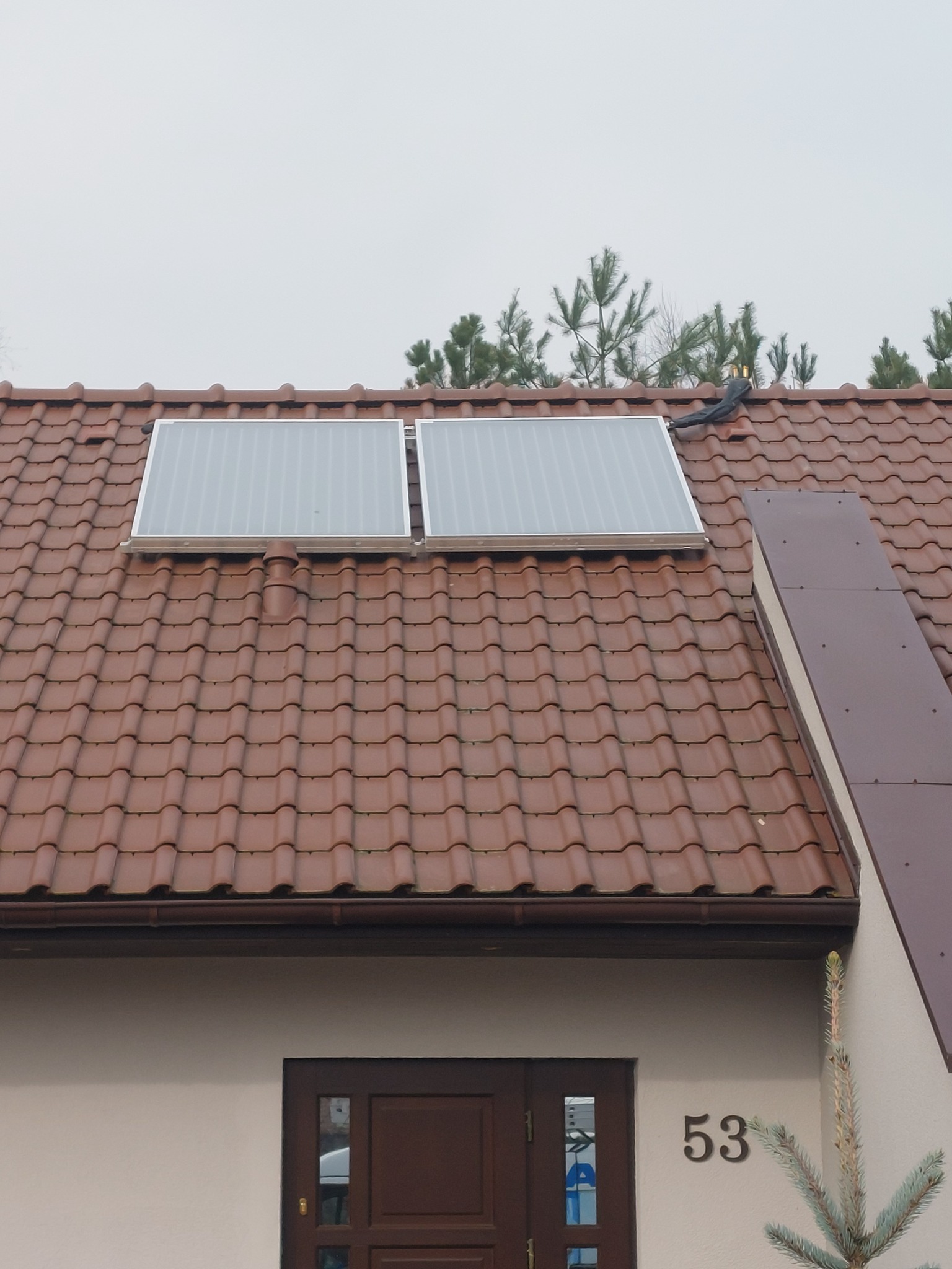 Dwa panele solarne na brązowym dachu domu jednorodzinnego z numerem 53, widoczne drzewa w tle, pochmurne niebo.
