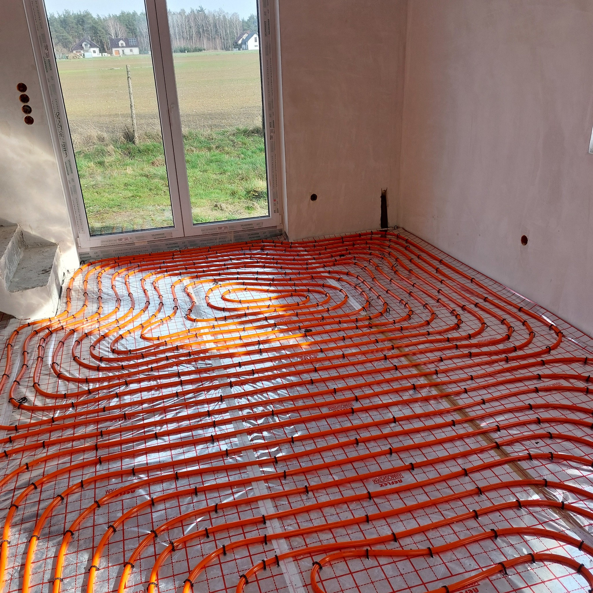 Pomarańczowe rury ogrzewania podłogowego ułożone spiralnie na folii z siatką w surowym wnętrzu, widok przez okno na pole i domy w oddali.