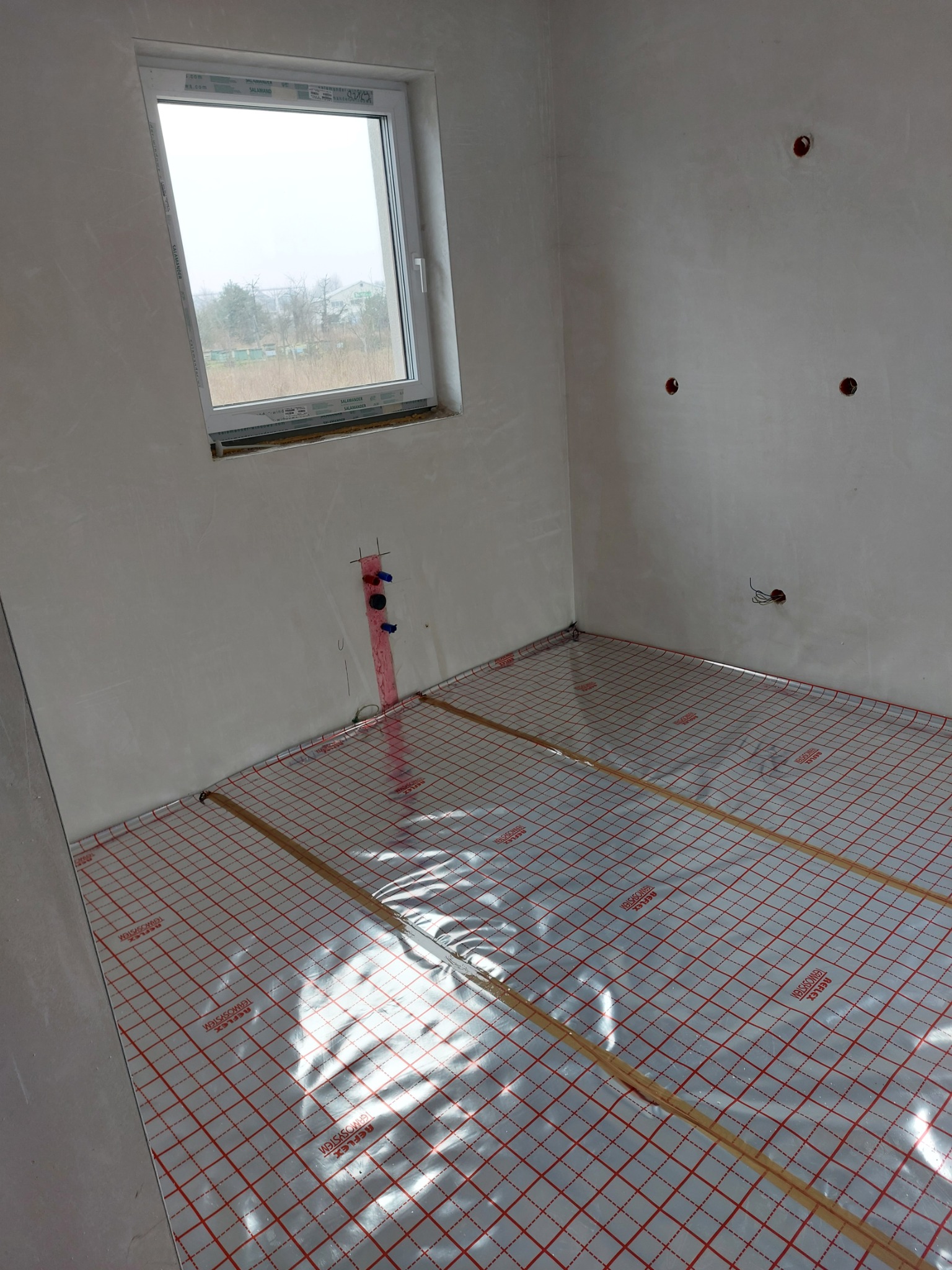 Przygotowanie do montażu ogrzewania podłogowego: mata grzewcza z folią aluminiową i czerwoną siatką na podłożu w surowym wnętrzu, widoczne wyprowadzenia instalacji wodnej i elektrycznej na ścianie.