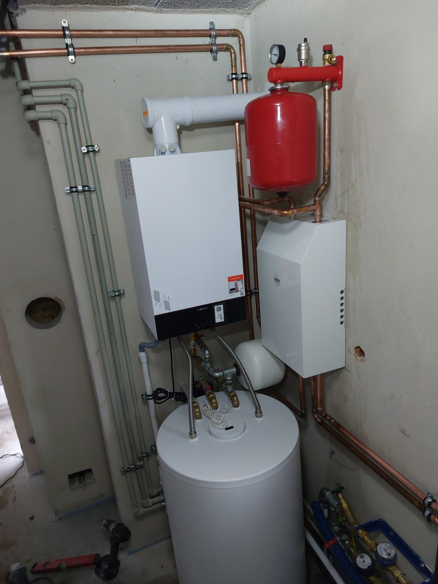 Nowoczesny, biały piec grzewczy z czerwonym zbiornikiem wyrównawczym i miedzianą instalacją w surowym pomieszczeniu, widoczny poziomica i wkrętarka na podłodze.