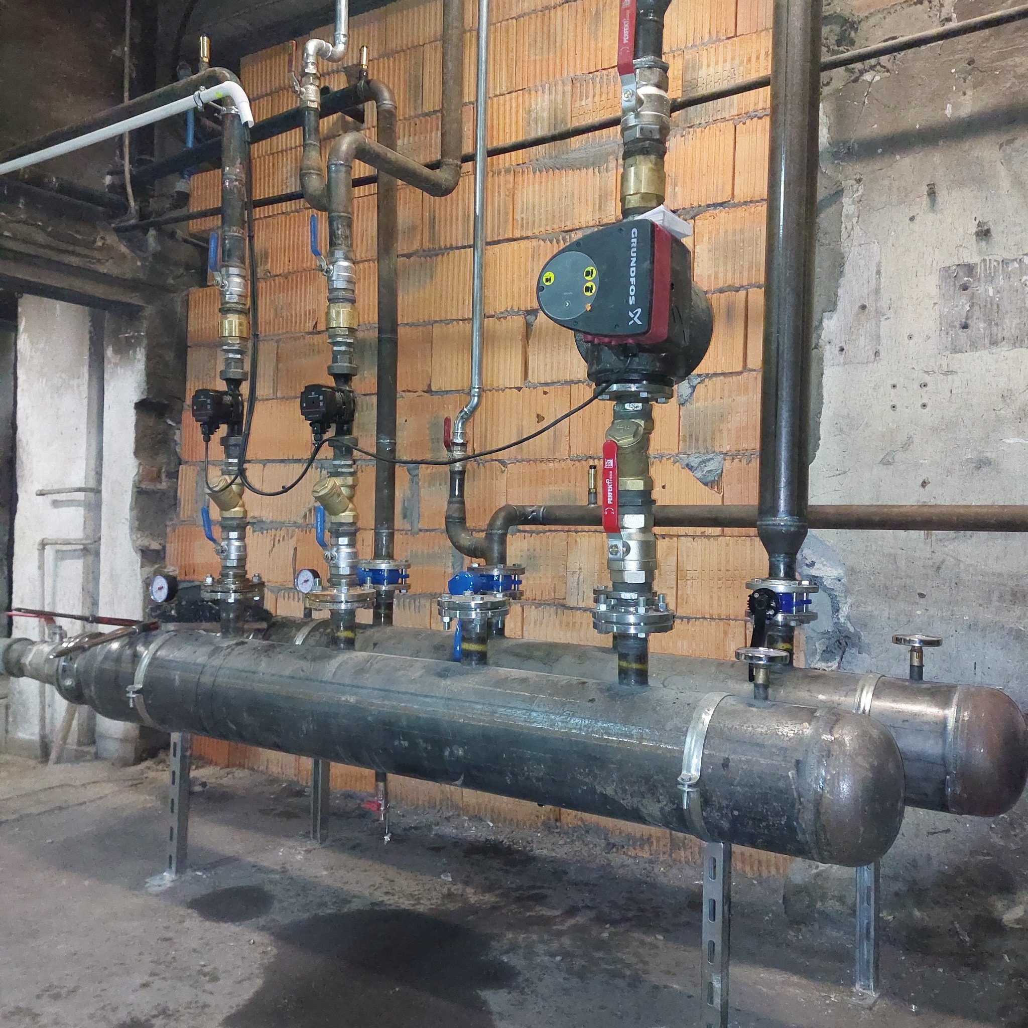 Rozbudowana instalacja grzewcza z pompą obiegową Grundfos, rurami stalowymi i miedzianymi, zaworami oraz manometrami, zamontowana przy ścianie z cegły dziurawki.
