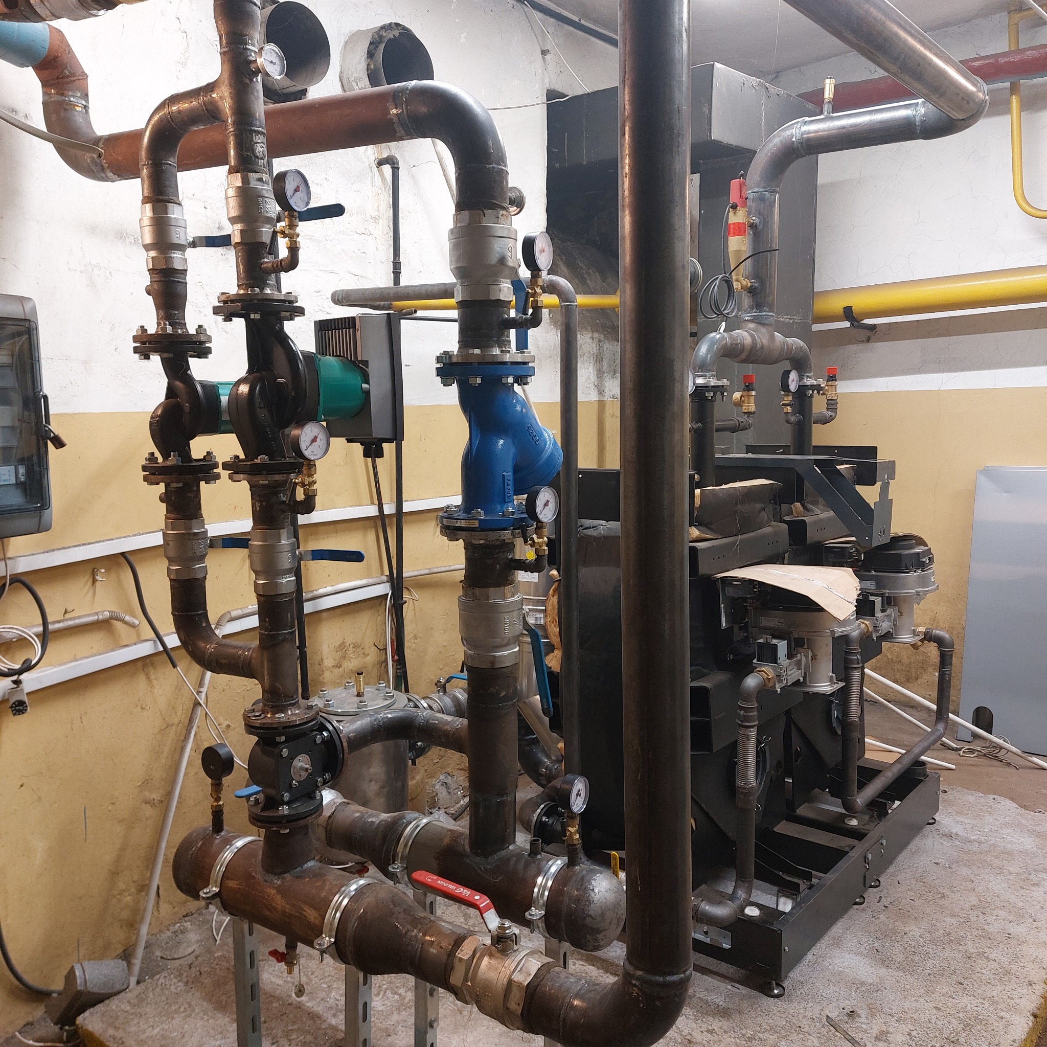 Złożona instalacja grzewcza z licznymi rurami, zaworami i manometrami w kotłowni w Wągrowcu, widoczne różne materiały i kolory rur.