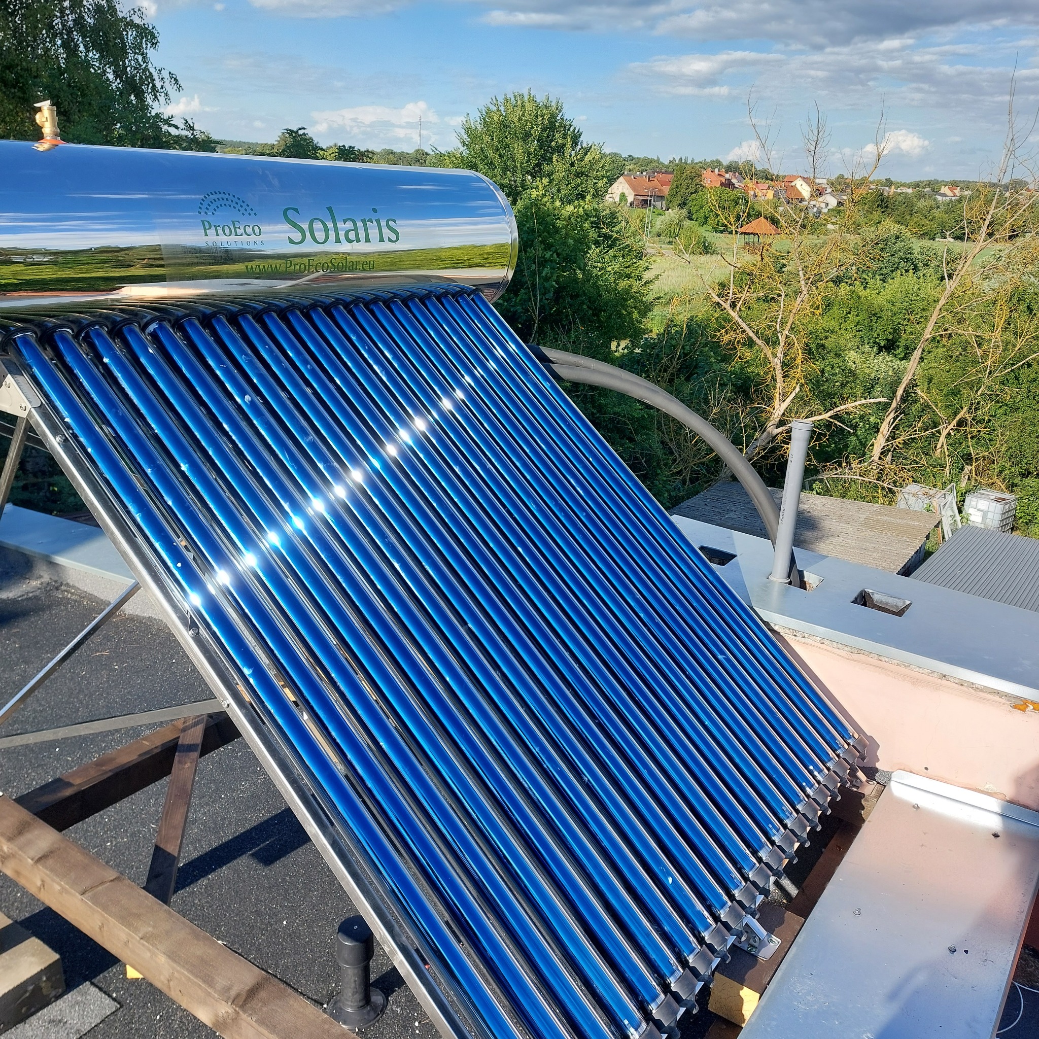 Kolektory słoneczne próżniowe z logo ProEco Solaris na dachu budynku, odbijające błękitne niebo i okoliczny krajobraz.