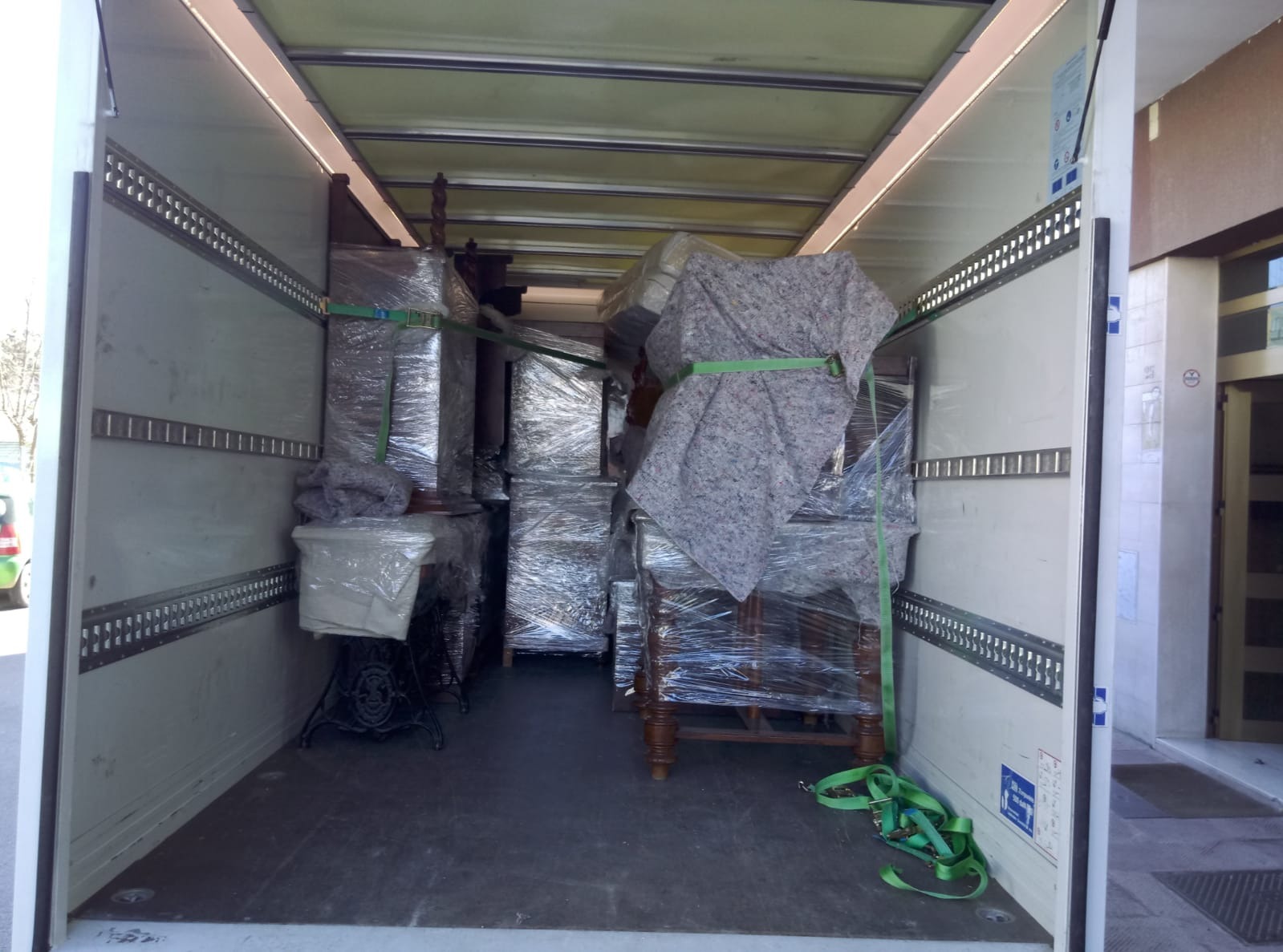 Wnętrze furgonetki załadowane meblami zabezpieczonymi folią i pasami transportowymi, widoczna stara maszyna do szycia z żeliwną podstawą.