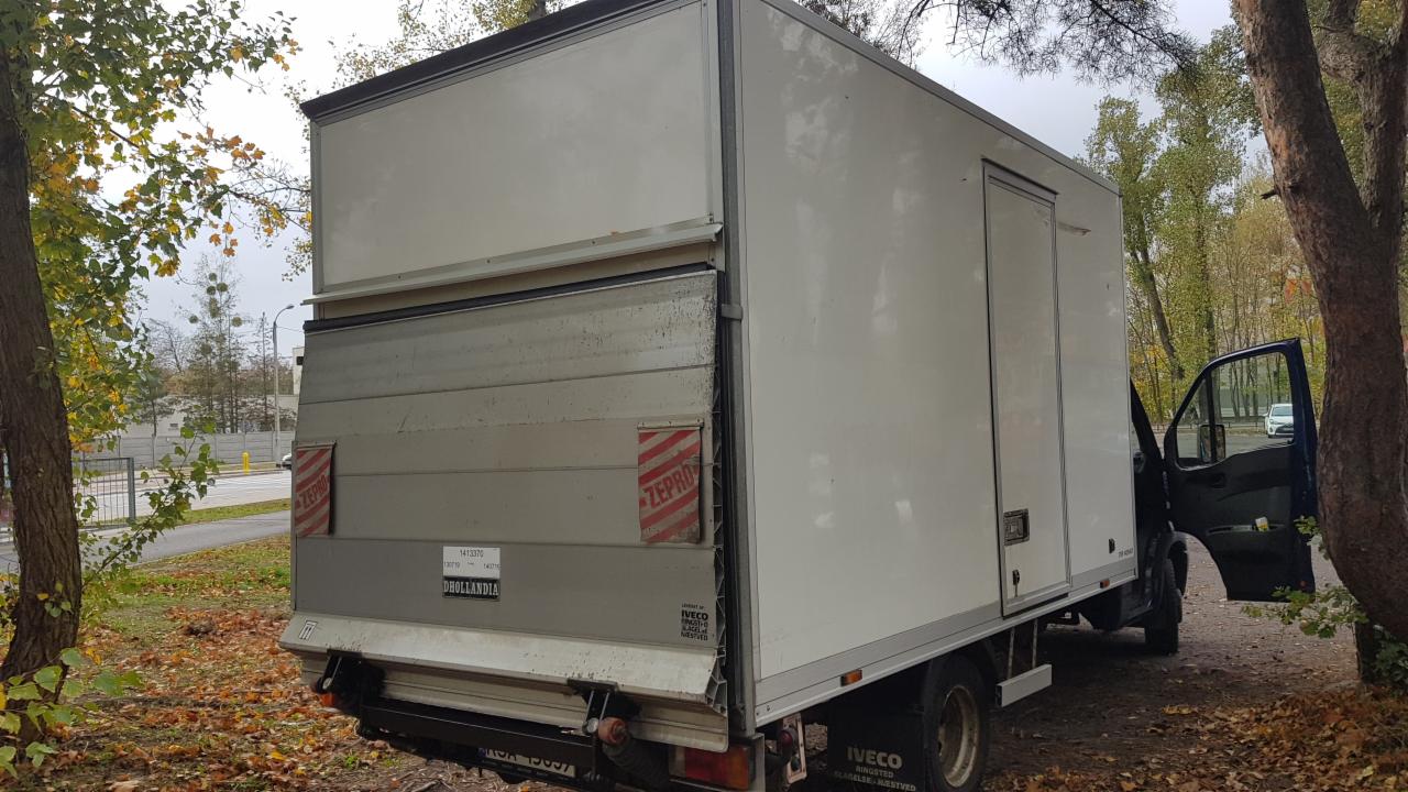 Biała furgonetka dostawcza Iveco z otwartymi drzwiami kabiny i częściowo otwartą tylną klapą, zaparkowana na ziemi pokrytej liśćmi w jesiennej scenerii.