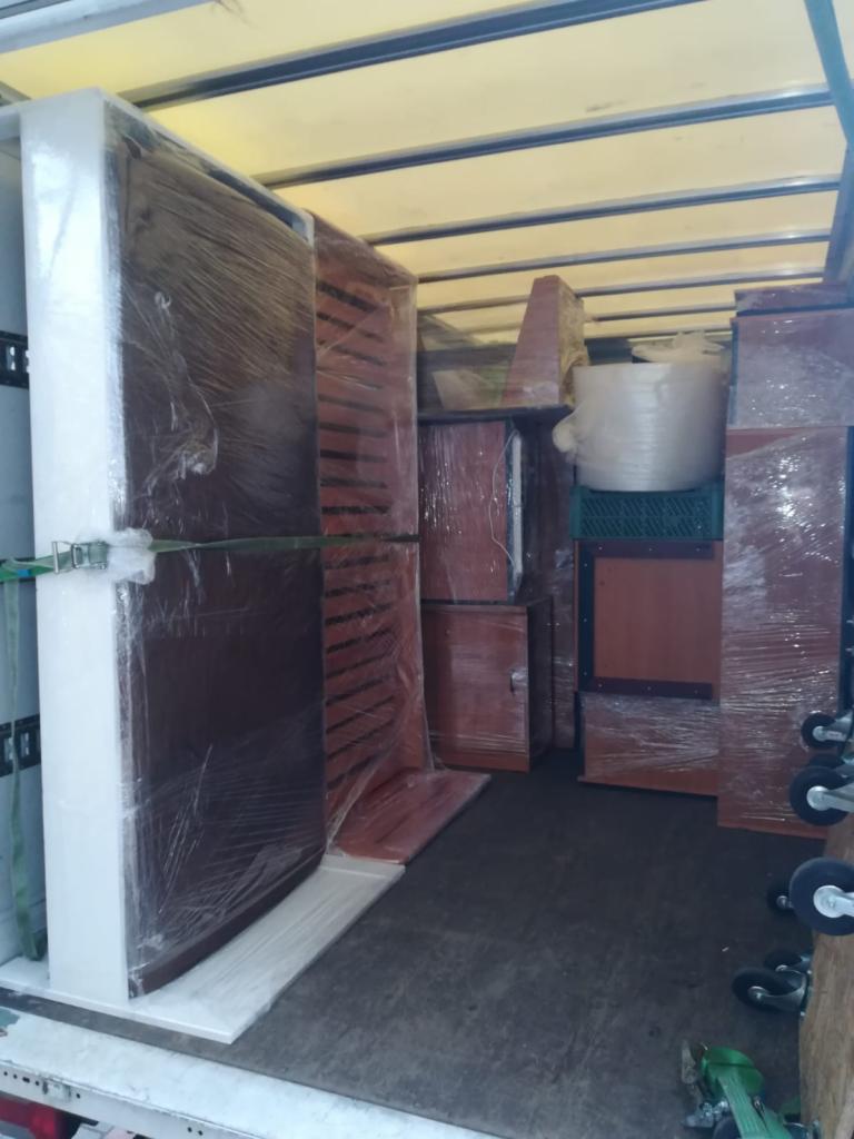 Wnętrze furgonetki załadowane meblami zabezpieczonymi folią stretch, przygotowanymi do transportu, widoczne wózki transportowe.