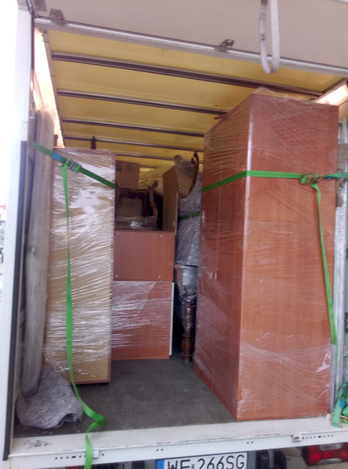 Wnętrze busa wypełnione meblami zabezpieczonymi folią i pasami transportowymi, widok z tyłu pojazdu, częściowo widoczna tablica rejestracyjna.