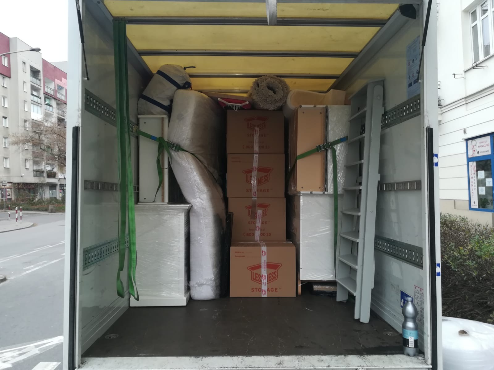 Wnętrze furgonetki wypełnionej spakowanymi meblami, kartonami z logo LESS MESS STORAGE i zabezpieczonej pasami transportowymi podczas przeprowadzki.