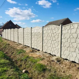 Długie, jasnoszare ogrodzenie z betonu imitującego kamień, biegnące wzdłuż działki z widocznymi domami w tle, świeżo obsypane ziemią.