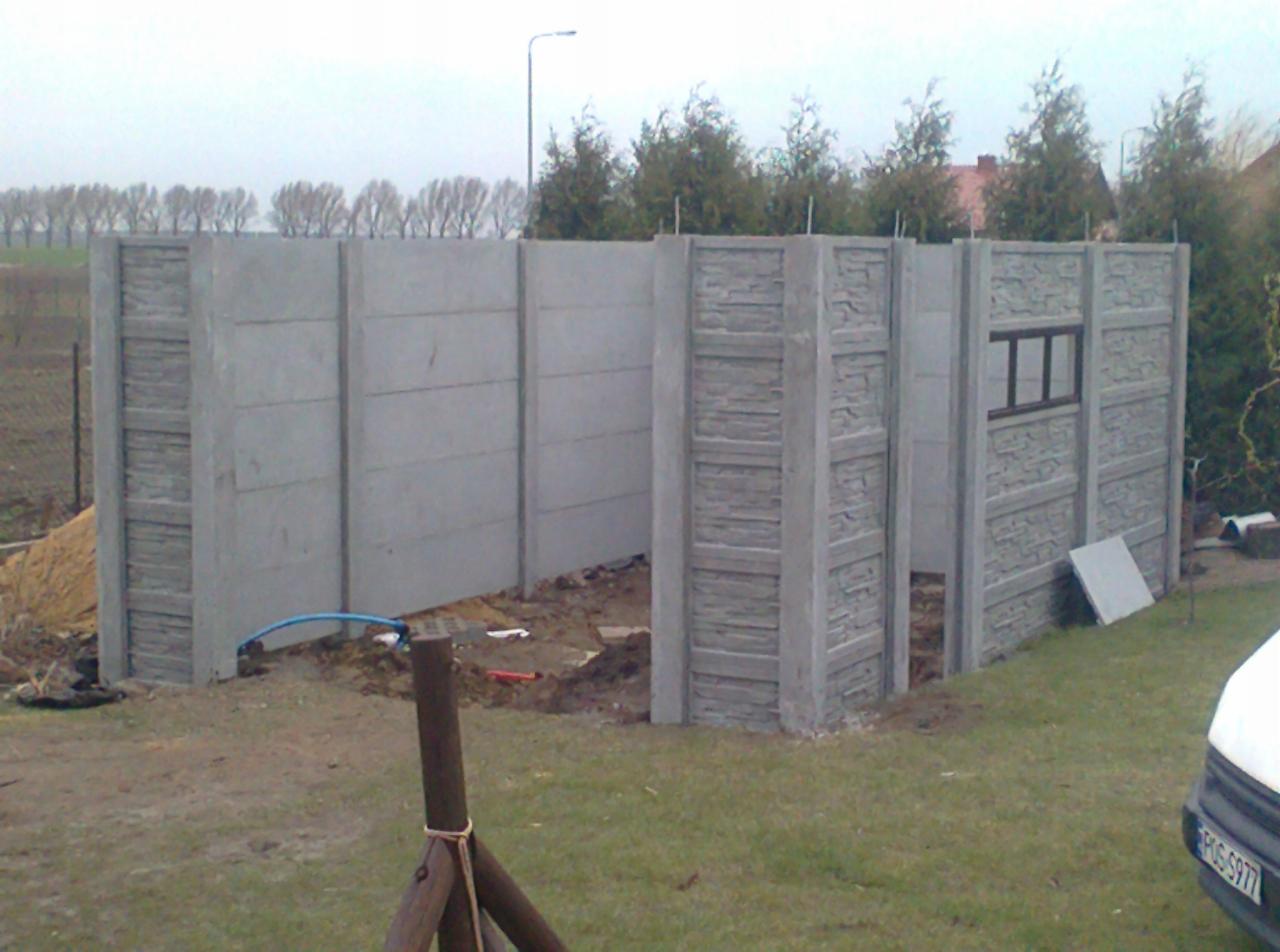 Montaż prefabrykowanego ogrodzenia z betonu architektonicznego, widoczne elementy w różnej fazie instalacji, fragment samochodu z widoczną tablicą rejestracyjną w prawym dolnym rogu.