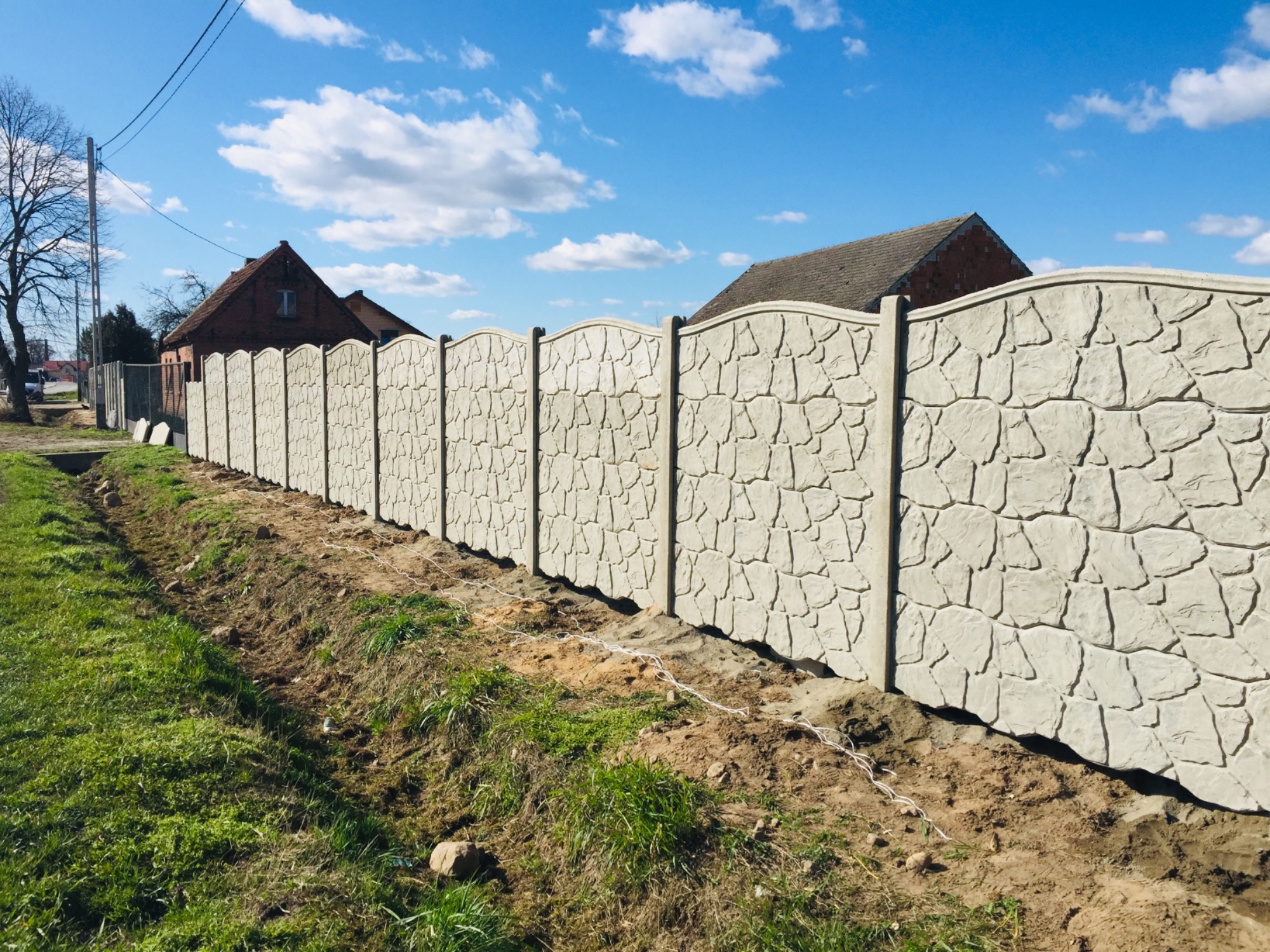 Długie, jasnoszare ogrodzenie z betonu imitującego kamień, biegnące wzdłuż działki z widocznymi domami w tle, świeżo obsypane ziemią.