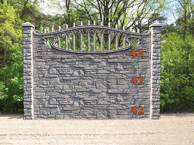 Fragment ogrodzenia z szarego kamienia łupanego z metalowym, ozdobnym wykończeniem u góry, na neutralnym tle z zielenią w tle.