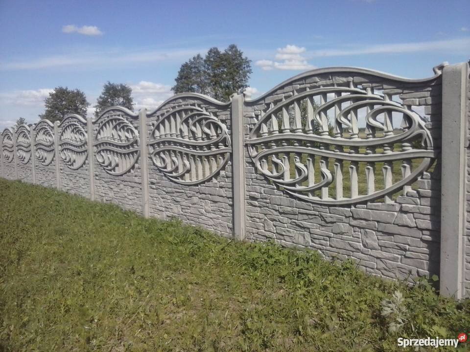 Długi, szary mur z prefabrykowanych elementów betonowych imitujących kamień, z dekoracyjnymi, ażurowymi przęsłami o falistym kształcie, na tle zielonej trawy i błękitnego nieba.