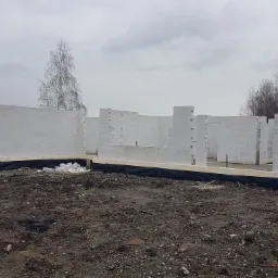 Wznoszenie ścian parteru domu z bloczków betonu komórkowego Ytong na fundamencie, widoczne zbrojenia i izolacja przeciwwilgociowa, w tle drzewa bez liści pod pochmurnym niebem.