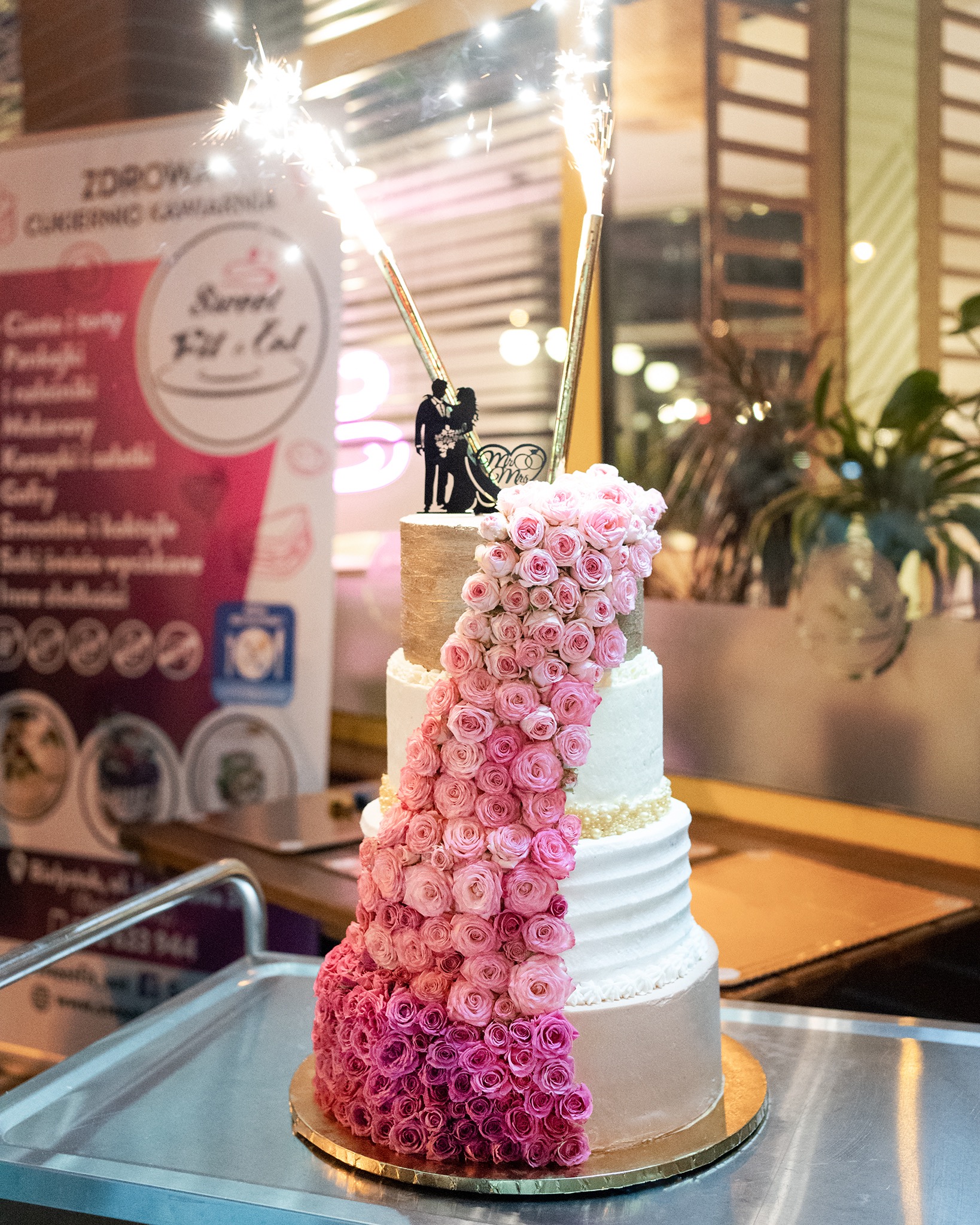 Wielopiętrowy tort weselny z kaskadą róż w odcieniach różu, zwieńczony figurką pary młodej i przymocowanymi racami.