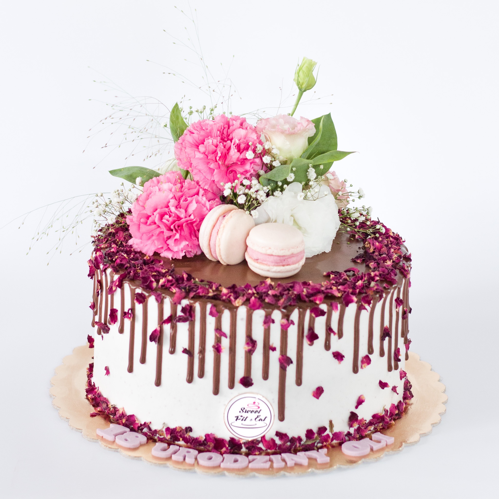 Tort urodzinowy z białym kremem, polewą czekoladową, dekoracją z płatków róż, kwiatów i makaroników na białym tle.