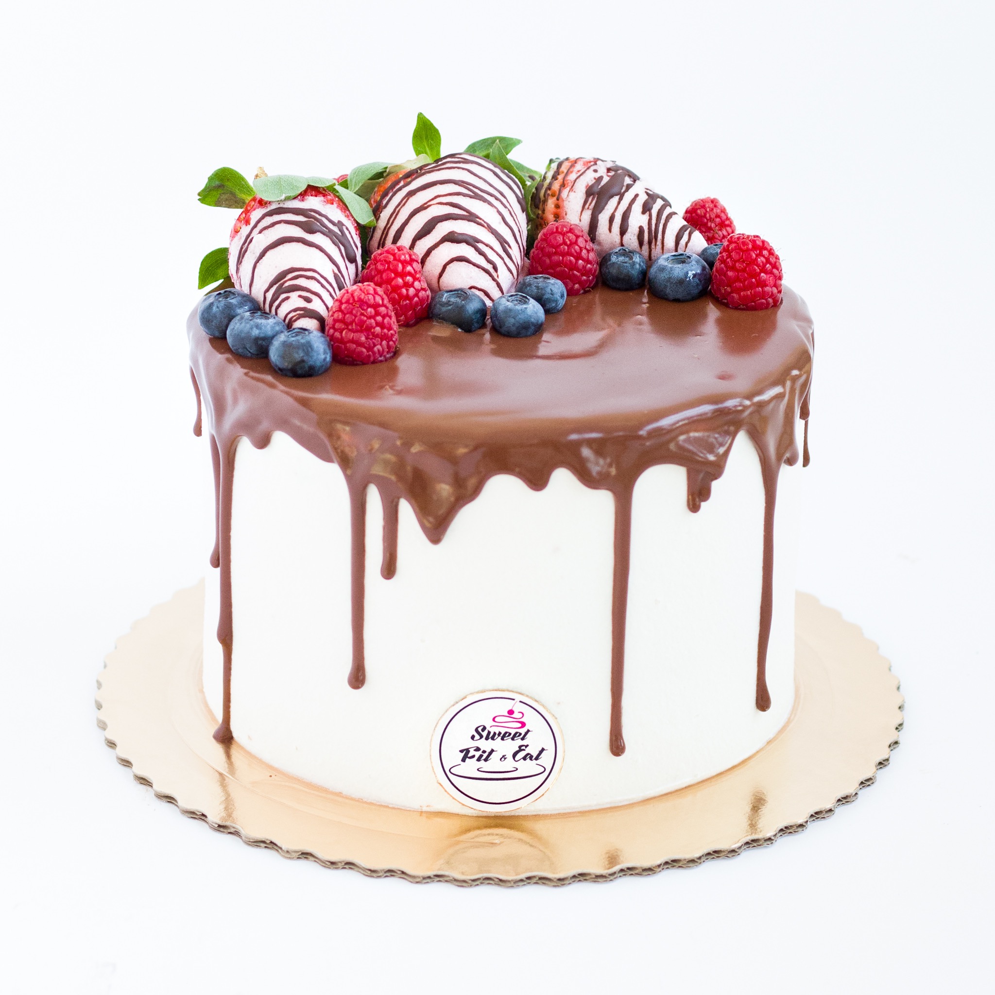 Tort z polewą czekoladową, udekorowany świeżymi truskawkami w polewie, malinami i borówkami, umieszczony na złotej podkładce.