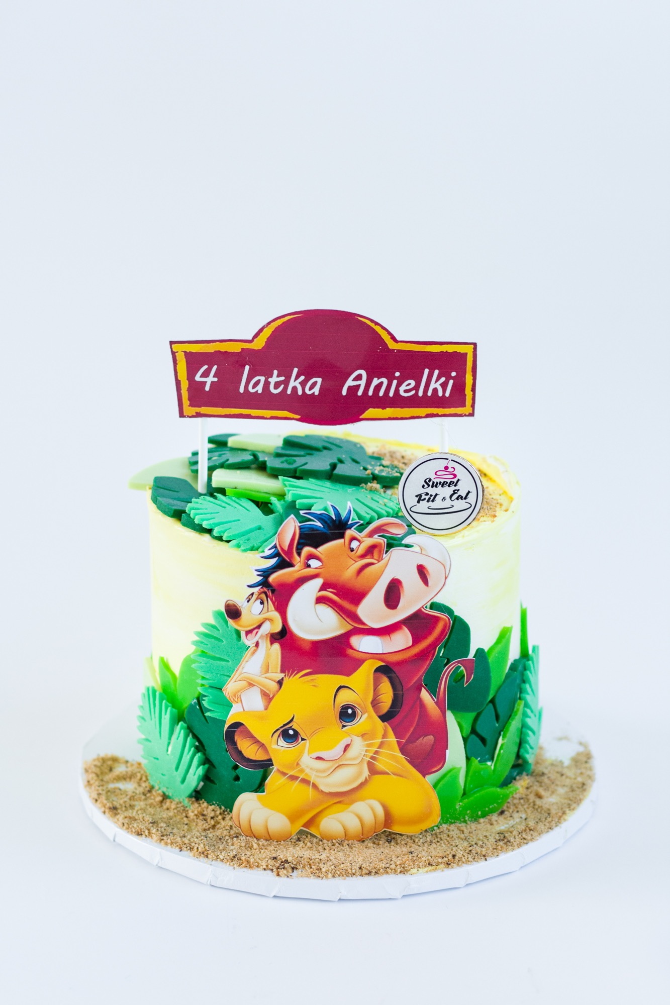 Tort urodzinowy z motywem Króla Lwa, dekorowany zielonymi liśćmi z lukru i topperem z napisem '4 latka Anielki', umieszczony na białym tle.