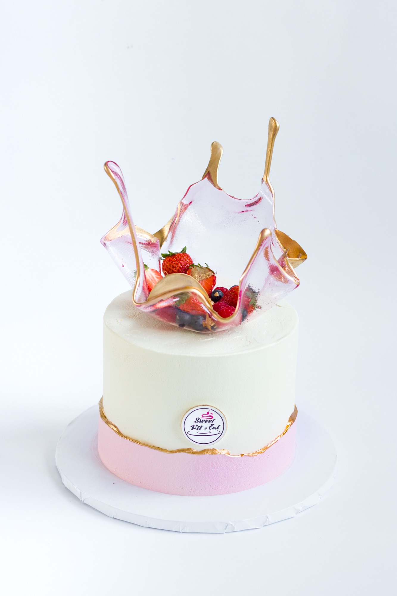 Dwupoziomowy tort z białą polewą, różowym akcentem i dekoracją z karmelu oraz świeżych owoców, na białym tle.