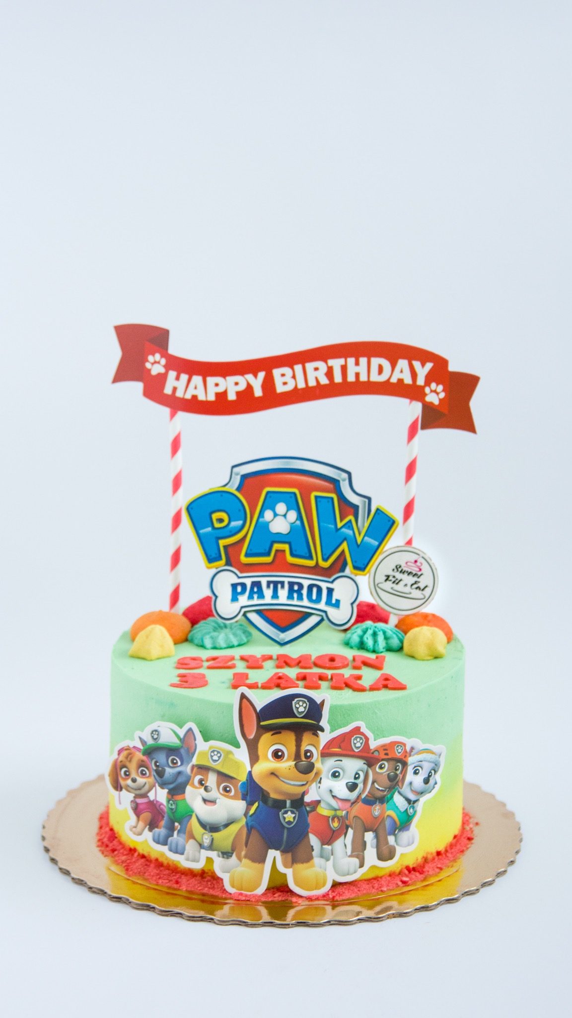 Tort urodzinowy z motywem Paw Patrol, z zielonym kremem, napisem 'Szymon 3 latka', dekoracjami z figurek psów i topperem 'Happy Birthday'.