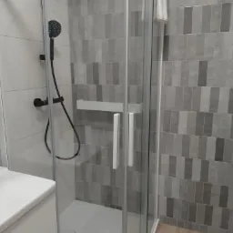 Nowoczesna kabina prysznicowa z czarną armaturą na tle szarych, prostokątnych płytek, z widoczną białą umywalką i ręcznikiem.