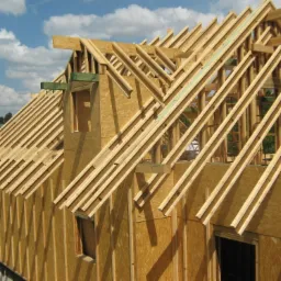 Konstrukcja domu w trakcie budowy: widoczne drewniane elementy szkieletu ścian i dachu, płyty OSB, niezadaszona bryła budynku na fundamencie w słoneczny dzień.