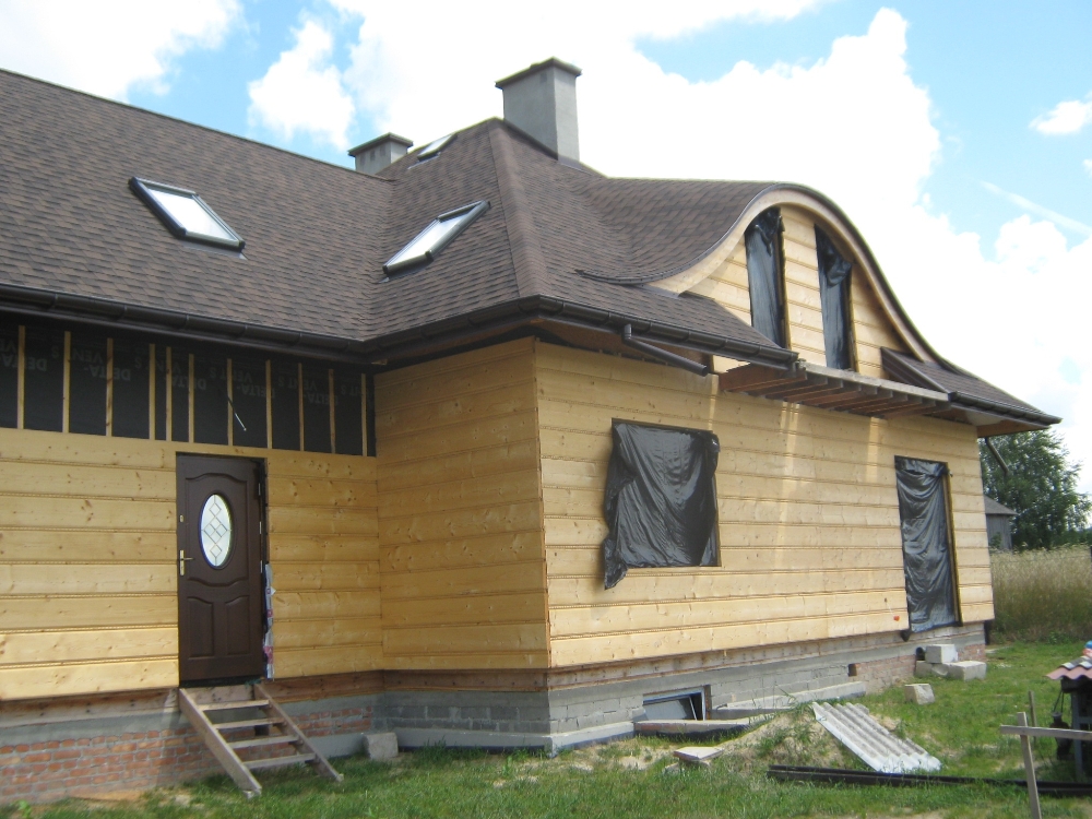 Dom w trakcie budowy z drewnianą elewacją, brązowym dachem z lukarnami i częściowo zakrytymi oknami folią ochronną, widoczne rusztowanie.