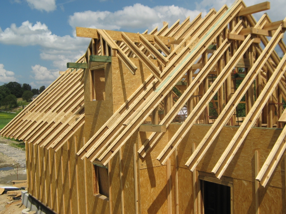 Konstrukcja domu w trakcie budowy: widoczne drewniane elementy szkieletu ścian i dachu, płyty OSB, niezadaszona bryła budynku na fundamencie w słoneczny dzień.