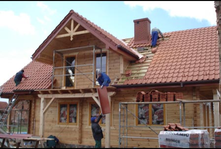 Budowa domu z bali klejonych: robotnicy układają czerwoną dachówkę na drewnianej konstrukcji, widoczne rusztowania i paleta dachówek firmy Koramic.