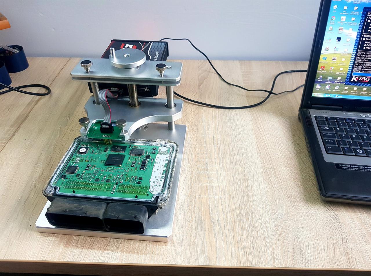 Elektroniczny moduł sterujący silnika (ECU) podłączony do urządzenia diagnostycznego na warsztatowym stole, obok laptopa z uruchomionym oprogramowaniem KTAG.