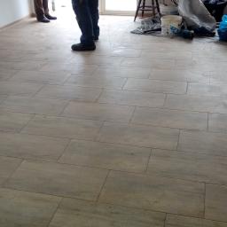 POL-BUD Paweł Wójcik - Ułożona podłoga z płytek imitujących drewno w trakcie prac wykończeniowych, widoczne narzędzia i materiały budowlane oraz fragmenty sylwetek dwóch osób.