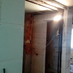 POL-BUD Paweł Wójcik - Montaż płyty karton gips na ścianie podczas remontu; widoczne surowe ściany, wystające przewody i fragment podłogi w stanie przed wykończeniem.