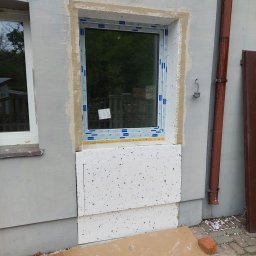 POL-BUD Paweł Wójcik - Montaż okna w ocieplanej ścianie zewnętrznej budynku; widoczne fragmenty styropianu, obróbka murarska wokół okna i zabezpieczenie kartonem na chodniku.