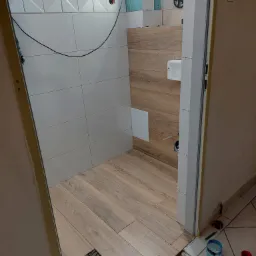 Układanie drewnopodobnych płytek podłogowych w łazience z częściowo wykończonymi ścianami w białych płytkach z dekoracyjnym pasem i imitacją drewna; widoczne krzyżyki dystansowe, kabel i narzędzia.