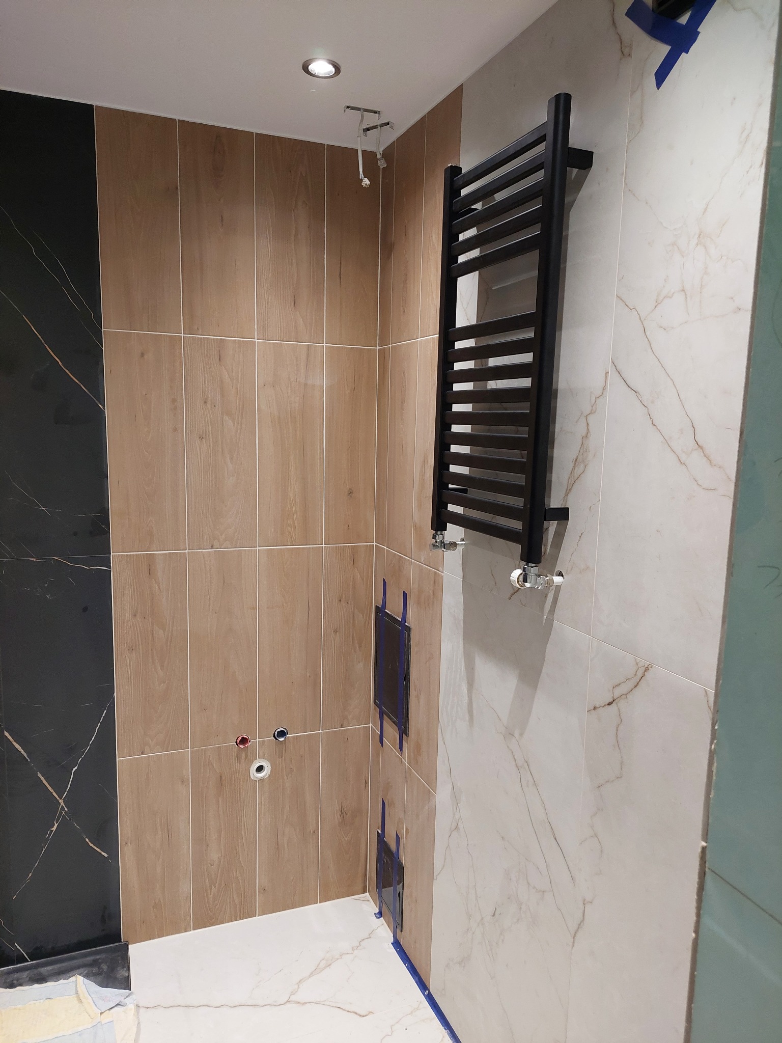 Częściowo wykończona łazienka z czarnym grzejnikiem drabinkowym na marmurowej ścianie i płytkami imitującymi drewno. Widoczne otwory na armaturę.