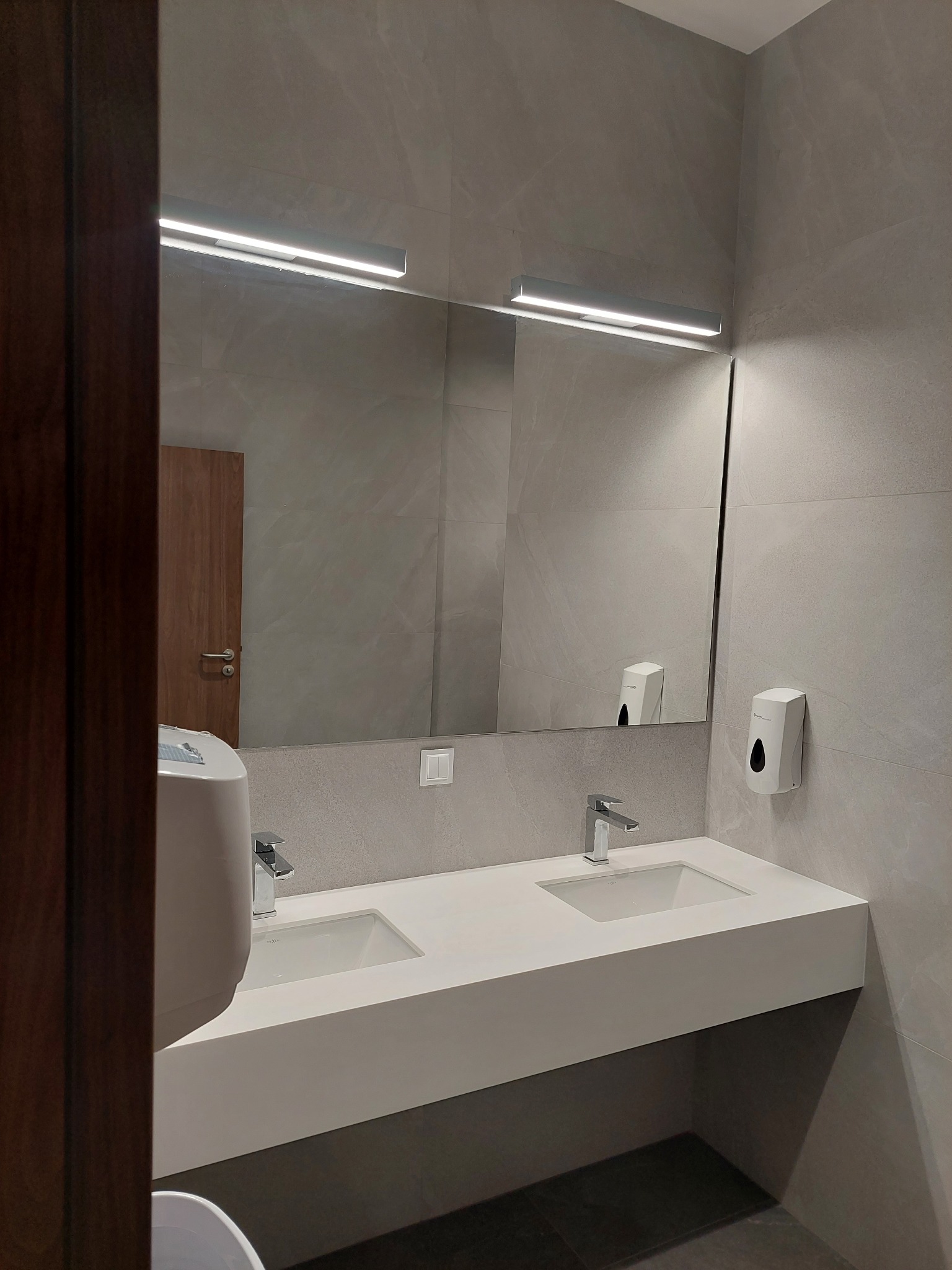 Nowoczesna łazienka z szarą płytką, podwójną białą umywalką i lustrem z oświetleniem LED. Minimalistyczny design, widoczny dozownik mydła i fragment drzwi.