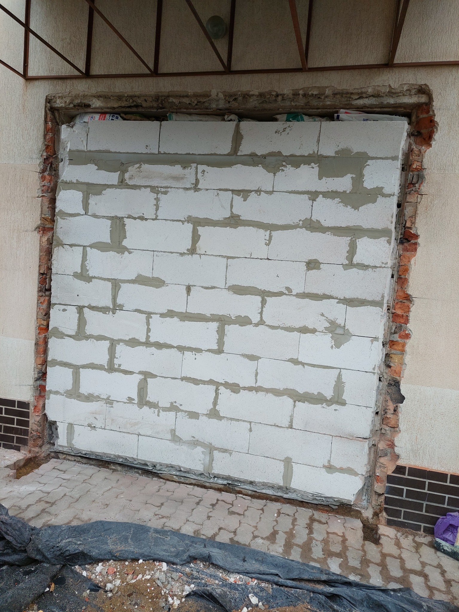 Zamknięty otwór okienny lub drzwiowy z wykorzystaniem białych bloczków betonowych, widoczne pozostałości starej ceglanej ramy otworu i fragment elewacji budynku.