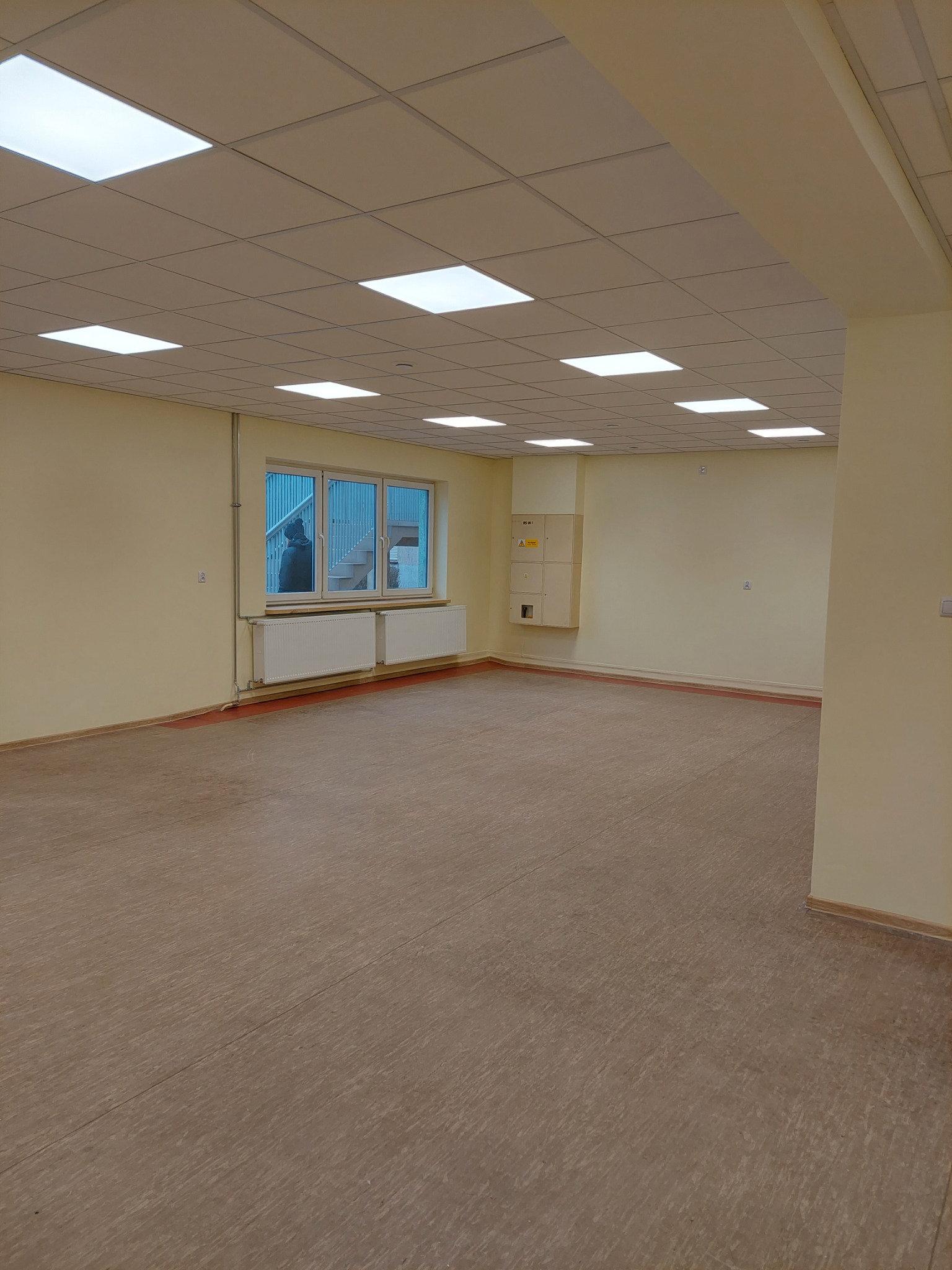 Puste pomieszczenie z beżowymi ścianami, brązową podłogą, oknem z widokiem na schody na zewnątrz i sufitem kasetonowym z kwadratowymi lampami