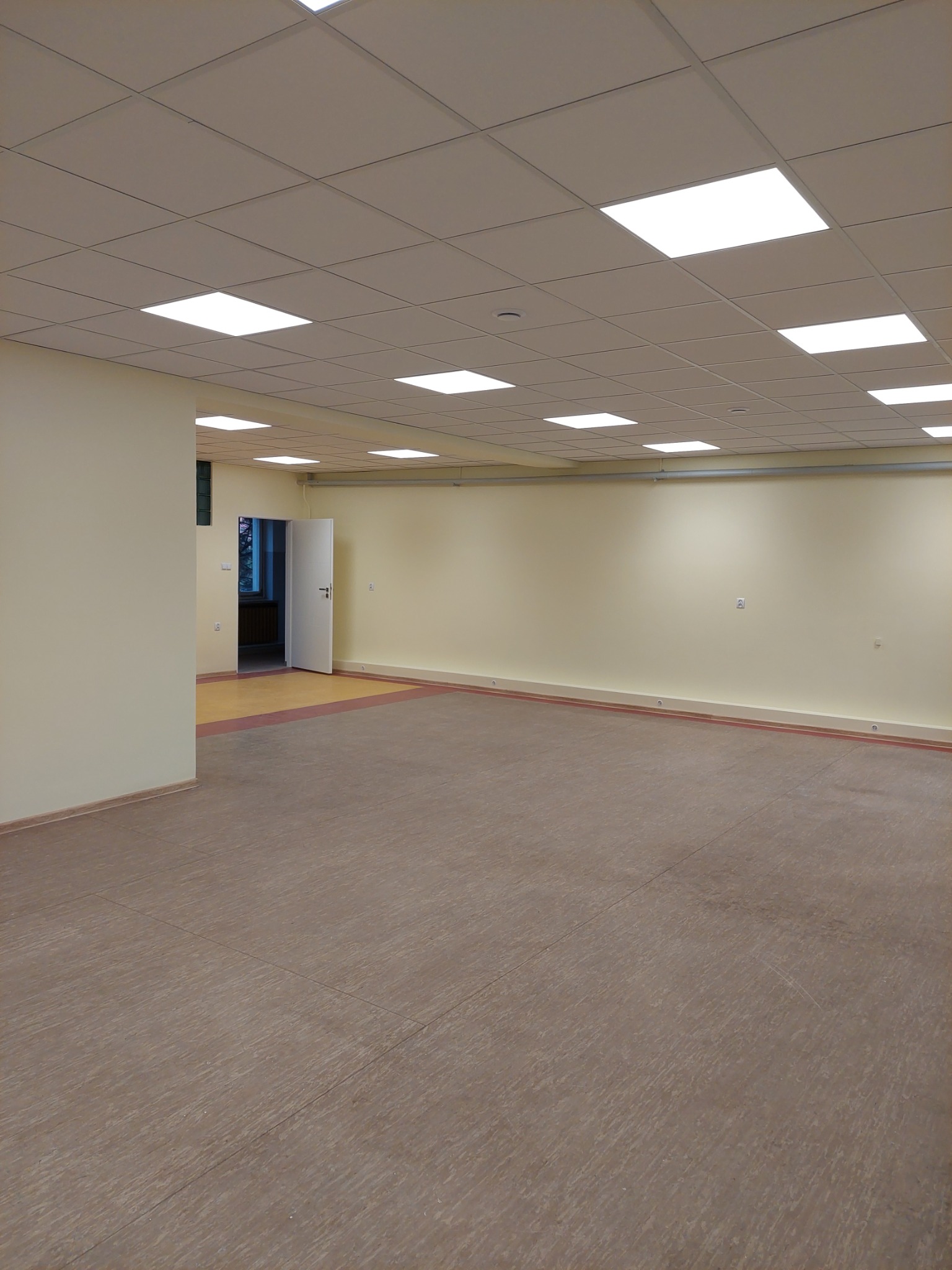 Pusta, jasna przestrzeń biurowa z panelami sufitowymi i świeżo położoną podłogą w odcieniach brązu. Otwarta biała drzwi w głębi ukazuje fragment innego pomieszczenia.