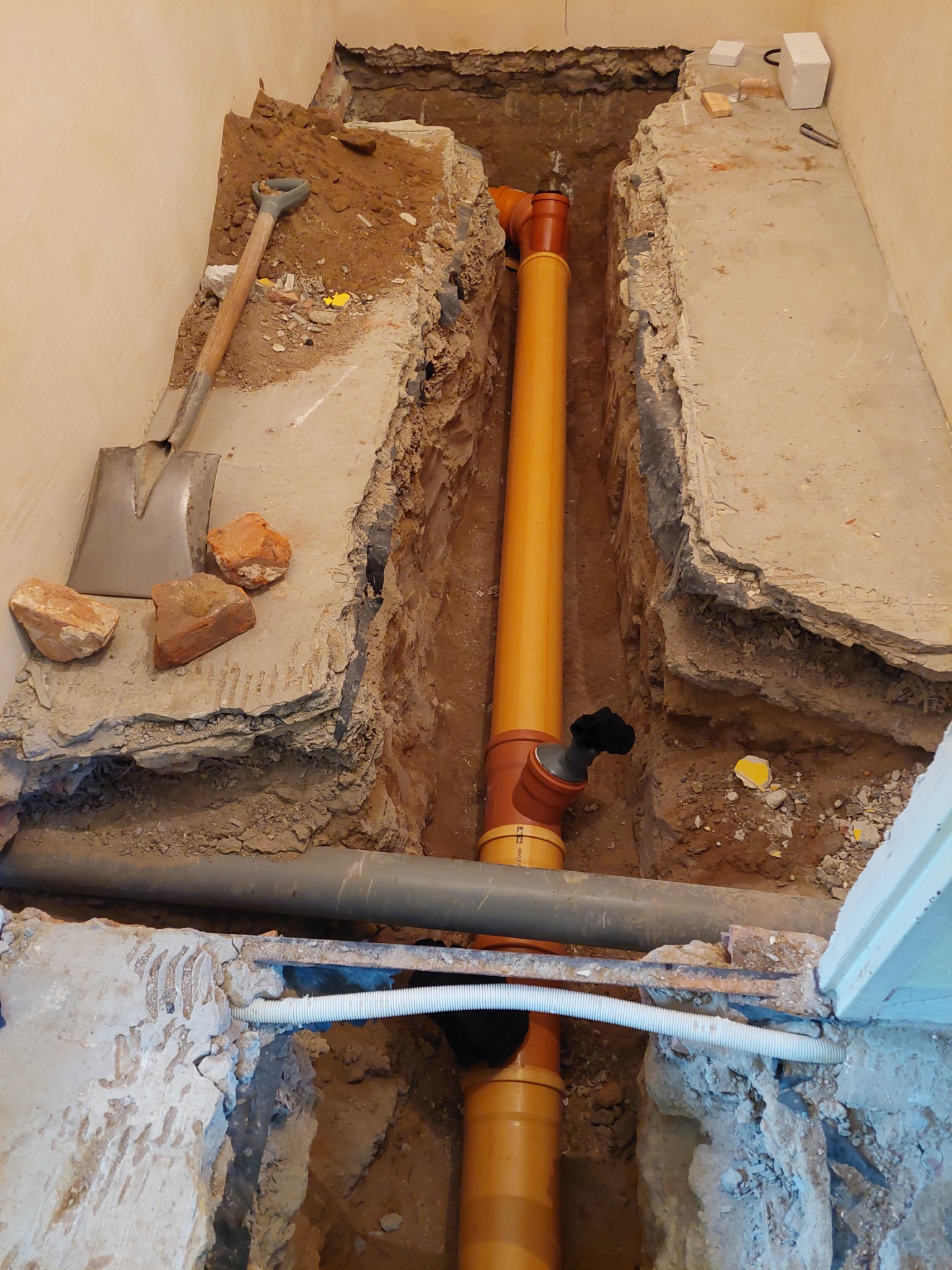 Wykop pod instalację kanalizacyjną z rurami w kolorze pomarańczowym, widoczny fragment ściany i podłogi w trakcie remontu łazienki w Głogowie, obok leży łopata.