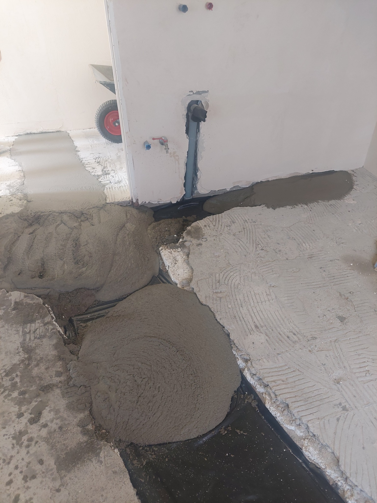 Przygotowanie podłoża w łazience: wylewka betonowa na folii hydroizolacyjnej, widoczne rury instalacji wodnej i fragment taczki w tle.