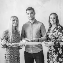 Trzy osoby prezentują portfolio z projektami wnętrz na tle jasnej ściany z subtelną fakturą.