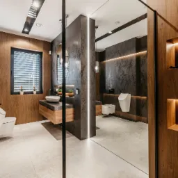 Nowoczesna łazienka z drewnianymi i grafitowymi elementami, podświetleniem LED, umywalką nablatową i wiszącą toaletą, widoczna przez szklaną ścianę.