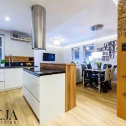 Nowoczesna kuchnia z wyspą i otwartą przestrzenią jadalną, białe szafki z czarnym blatem, drewniane akcenty, dekoracyjna okleina drzwi z motywem roślinnym, oświetlenie punktowe i wiszące lampy.