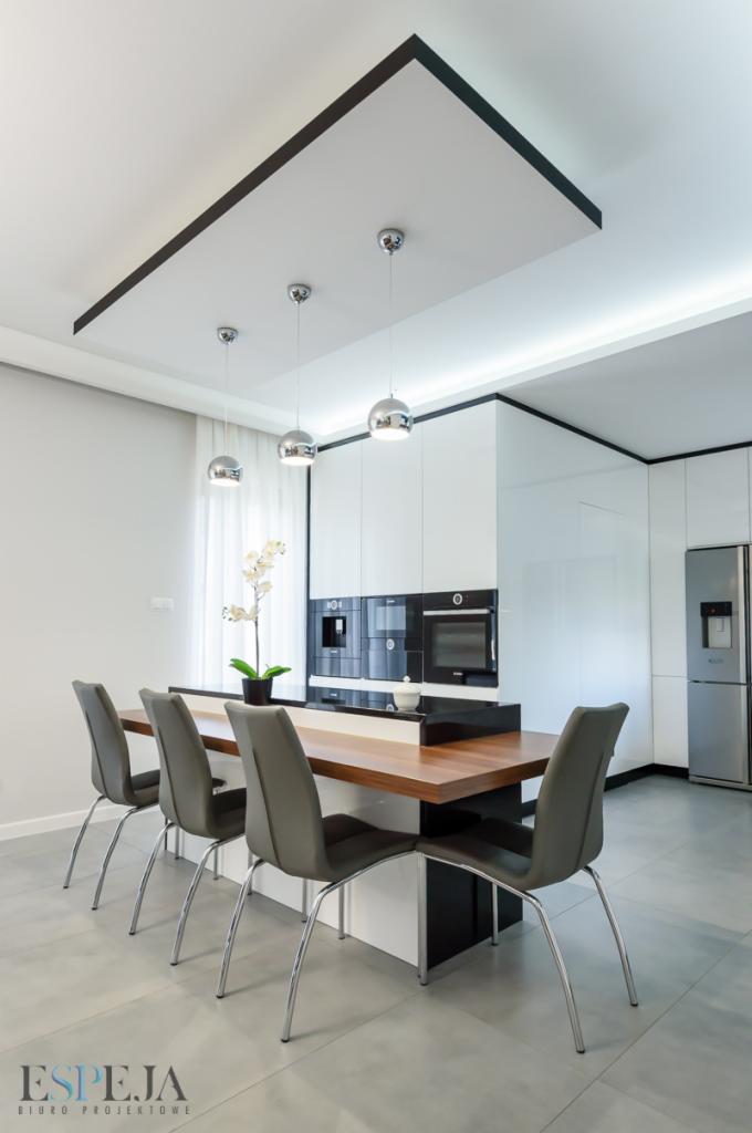 Nowoczesna kuchnia z wyspą i czterema krzesłami, oświetlona trzema okrągłymi lampami wiszącymi, z zabudową kuchenną w bieli i czerni oraz stalową lodówką.