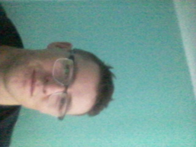 Portret mężczyzny w okularach na tle turkusowej ściany z widoczną częścią ramienia.