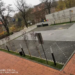 Widok z góry na nowo wykończony teren z nawierzchnią z drobnego żwiru, oświetlony lampami, z młodymi drzewkami wzdłuż krawędzi i z elementami z kostki brukowej w tle.