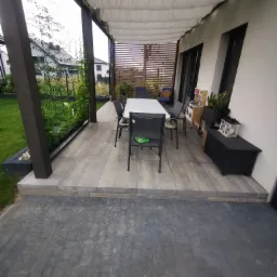 Zadaszony taras z szarych płyt, wyposażony w stół i krzesła, z zielonymi roślinami pnącymi się po pergoli i skrzynią na poduszki w rogu.