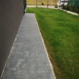 Szara ścieżka z drobnej kostki prowadząca wzdłuż ciemnej ściany budynku, obok trawnik z widoczną siatką ogrodzeniową w tle.